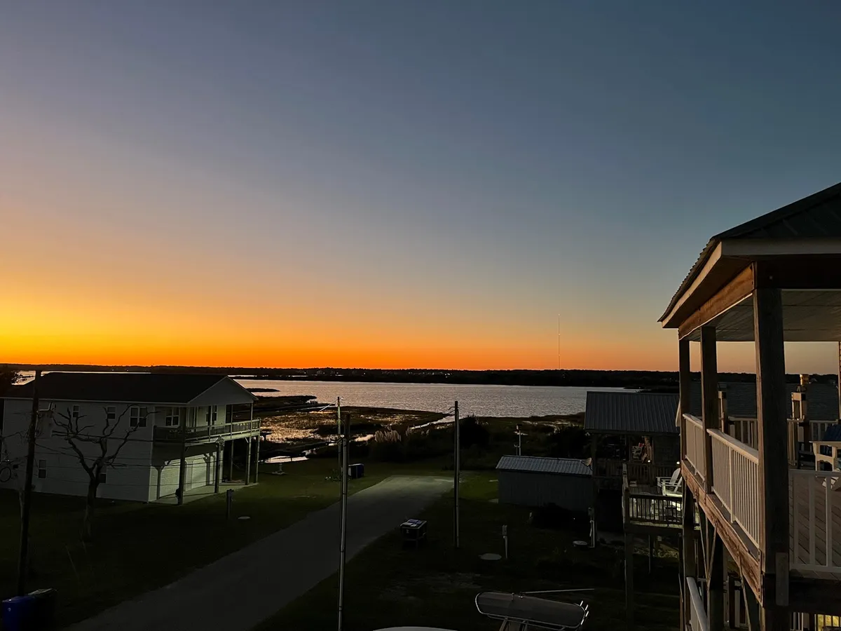 A tranquil view of the sun setting over the water, with the silhouette of buildings in the distance and a pathway leading directly to the water's edge. The scene is bathed in warm evening light, evoking a sense of peacefulness and connection to the waterfront surroundings.
