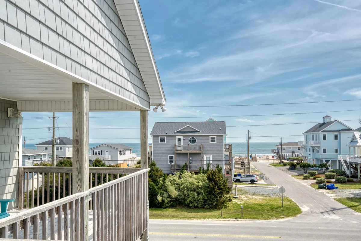 The image showcases a stunning view overlooking a quiet street lined with beach houses, with the sparkling ocean just one block away. The hero shot emphasizes a spacious balcony perfect for sunset watching, while the tranquil neighborhood and inviting sands evoke a sense of relaxation and family fun.