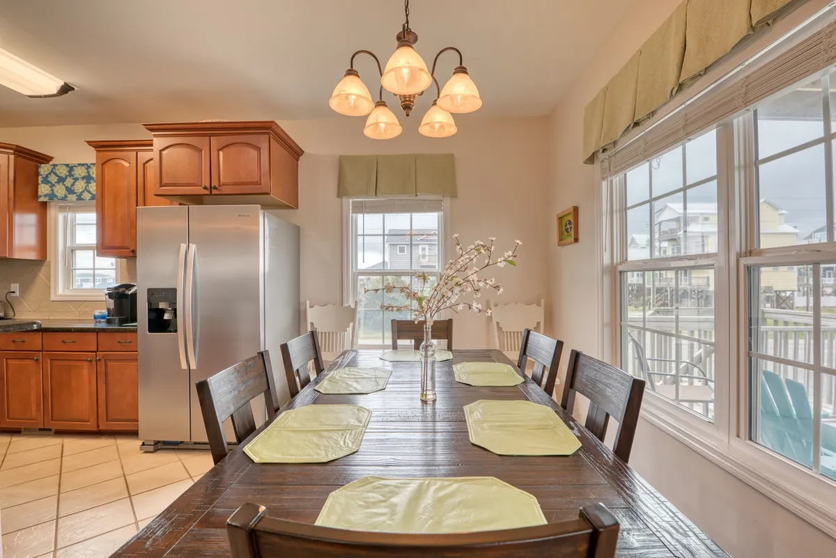 A cozy dining area with a large table set for gatherings, illuminated by a soft light fixture. Sunlight streams in through multiple windows, while the kitchen cabinets are visible in the background. The space is designed for shared meals, morning rituals, and celebrations with loved ones.