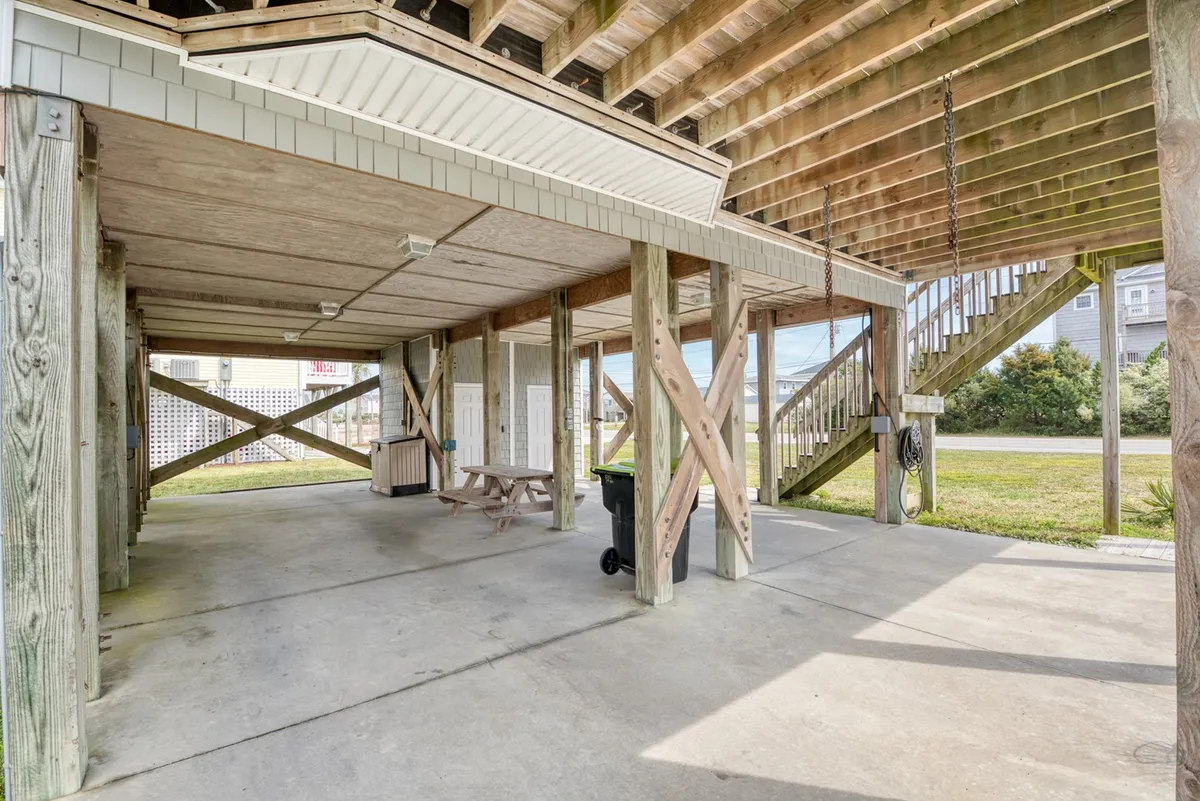 The image displays a covered parking area beneath the house, supported by sturdy wooden pillars, with a picnic table and a staircase leading up to the main living area. The space is generous, providing ample room for vehicles and potentially a boat trailer.