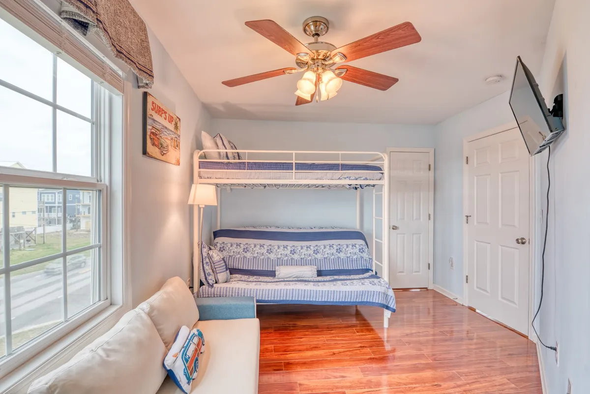 A cozy kids' bunk room featuring a bunk bed (twin over queen), a sofa, a flat-screen TV, a large window, and wooden floors. The space is playful and welcoming, perfect for sleepovers, movie nights, and creative fun.