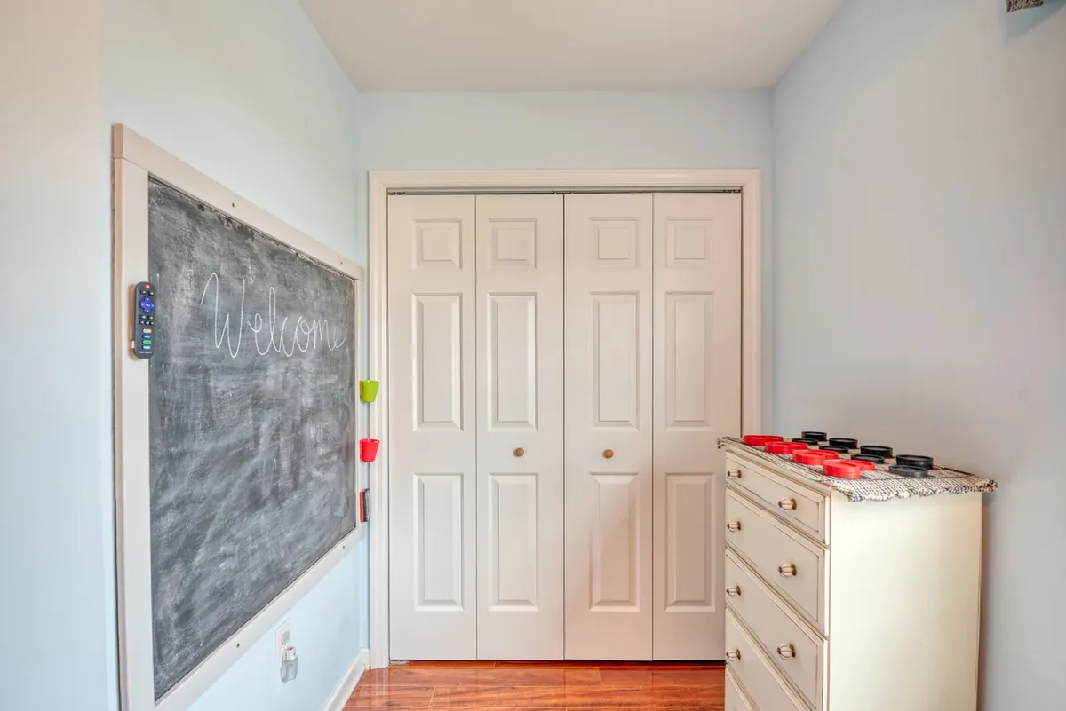 A cheerful room with a 'Welcome' chalkboard, white closet doors, and a chest of drawers adorned with colorful items. The playful, inviting decor suggests a family-friendly environment with flexible spaces for personal belongings and creative activities.