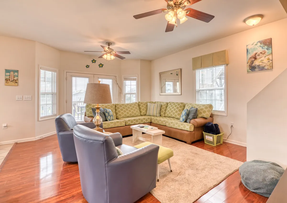 A bright living area featuring a green sectional sofa, a coffee table, and decorative touches like a lamp and framed artwork. The room is welcoming, ideal for relaxing or gathering as a group.