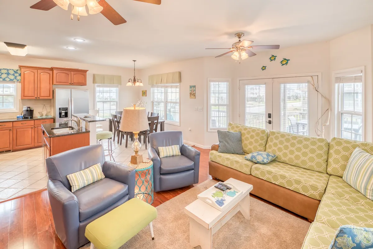 The image shows a spacious, cheerful great room featuring a green sectional sofa and blue armchairs, with a view into the adjacent kitchen area and a dining table. The open layout invites family gatherings, relaxation, and shared meals, with natural light streaming in.