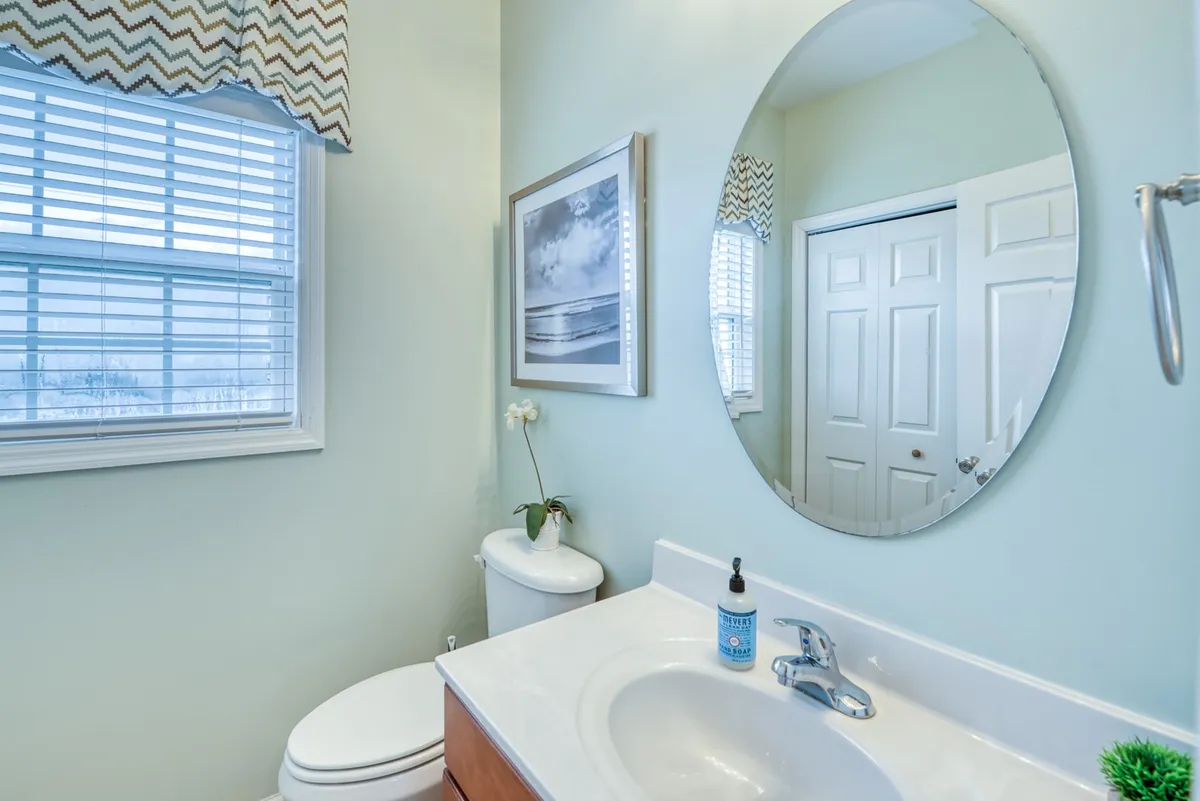 A bright, inviting bathroom with a round mirror above a sink, a faucet, a framed beach photo, and a window with blinds. The space feels fresh and rejuvenating, suggesting a place to rinse off after time spent outdoors.